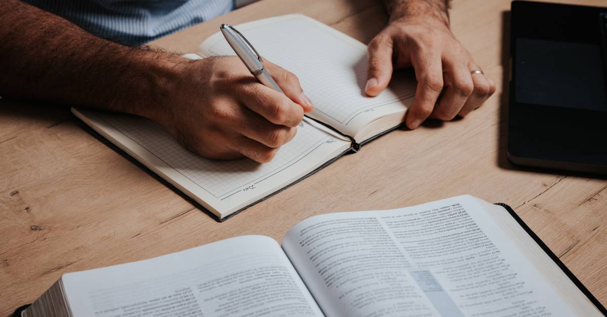 A man writing notes on a fresh page in his notebook. An open Bible lays on the table in front of him.