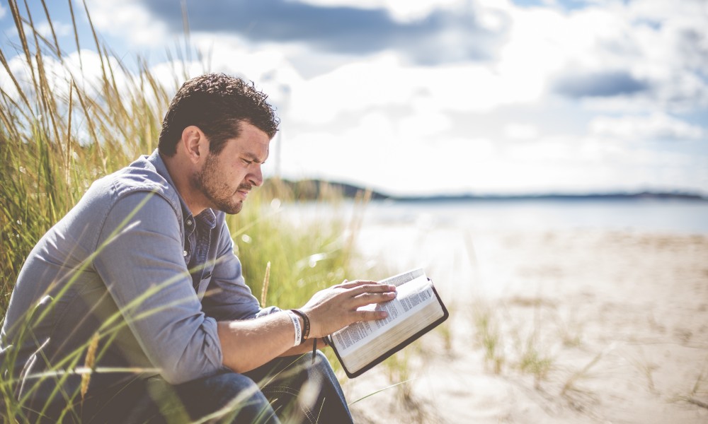 Man wearing a faded denim shirt and a few bracelets seated on a low sand dune on a beach reading a Bible
