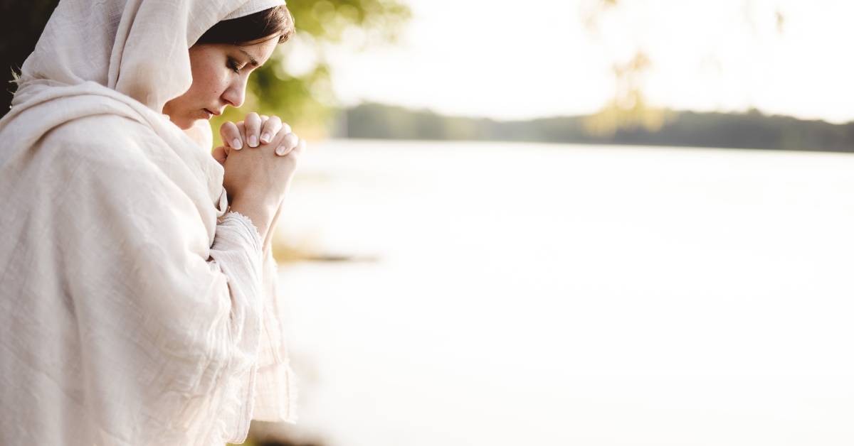 A woman wearing a cream-colored linen robe and head covering bowing her head in prayer by a body of water.