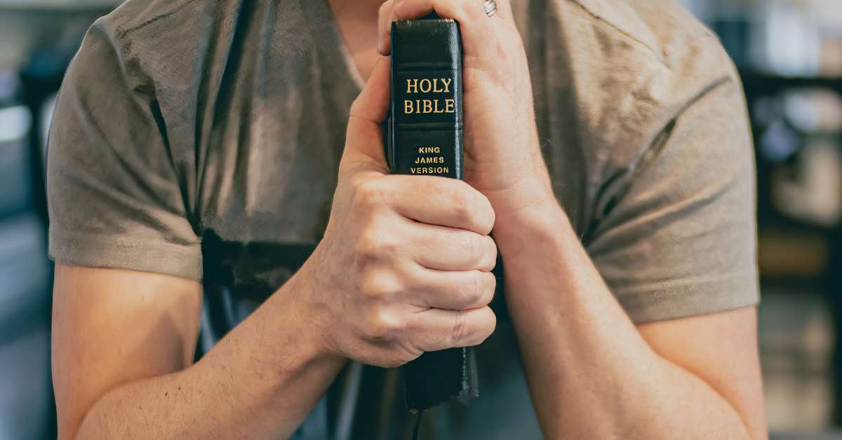 A man sitting with his elbows on a table clutches a black leather Bible. The Bible's spine is labeled the King James Version.