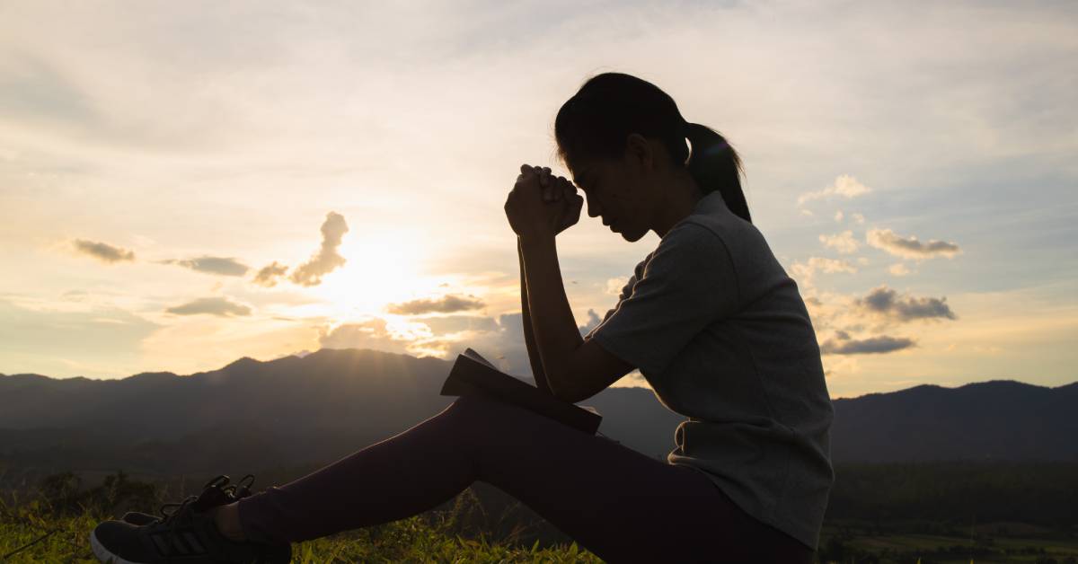 Silhouette of a person sitting outdoors and praying with a book on their lap. The sun is behind mountains on the horizon.