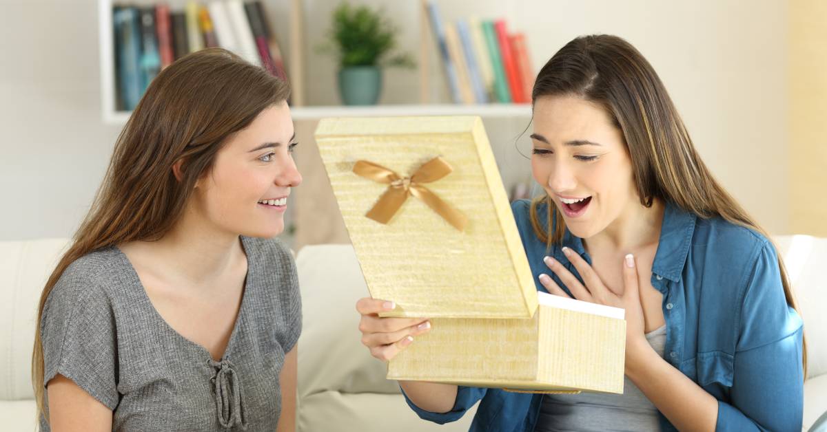 A woman sitting on a couch opening her mouth in surprise as she looks in a gift box. Another woman sitting beside her smiles.