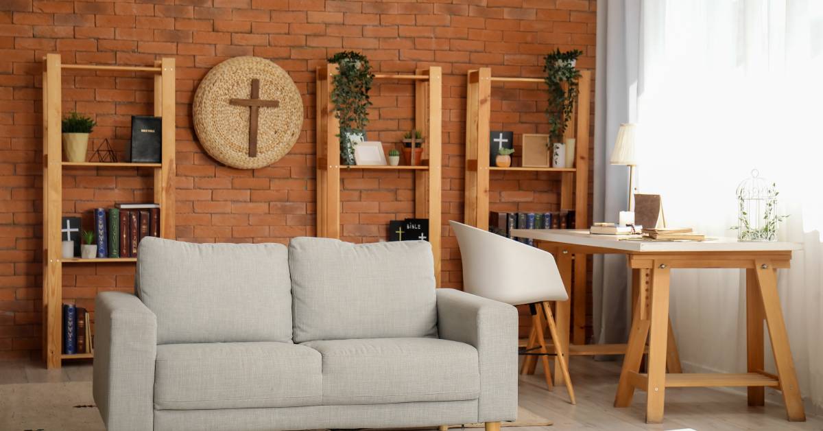 A white and beige living room with a red brick wall. A large Christian cross art piece hangs on the wall between bookshelves.