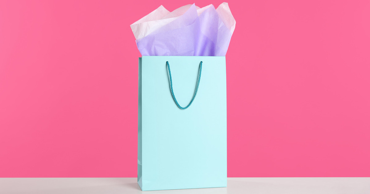 A pastel turquoise gift bag is on a table against a pink background. Purple and white tissue paper pops out of the bag opening.