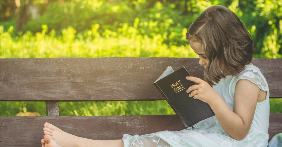 A child sitting on a wooden bench and reading the Bible. Sunshine is illuminating the tall green grass behind the bench.