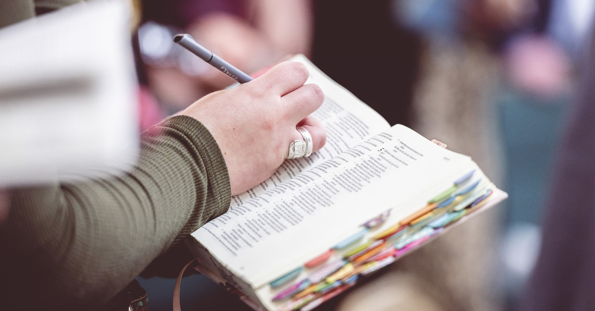 A woman at a church service takes notes in a wide-margin Bible she holds in her hand. The Bible has colorful indexing tabs.